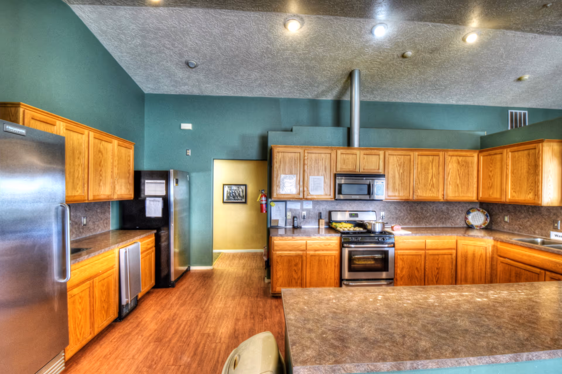 A spacious kitchen with wooden cabinets, stainless steel refrigerator, stove, microwave, and dishwasher. The walls are painted teal, and the floor has a wood finish. The kitchen has a large countertop island in the foreground and recessed lighting in the ceiling.
