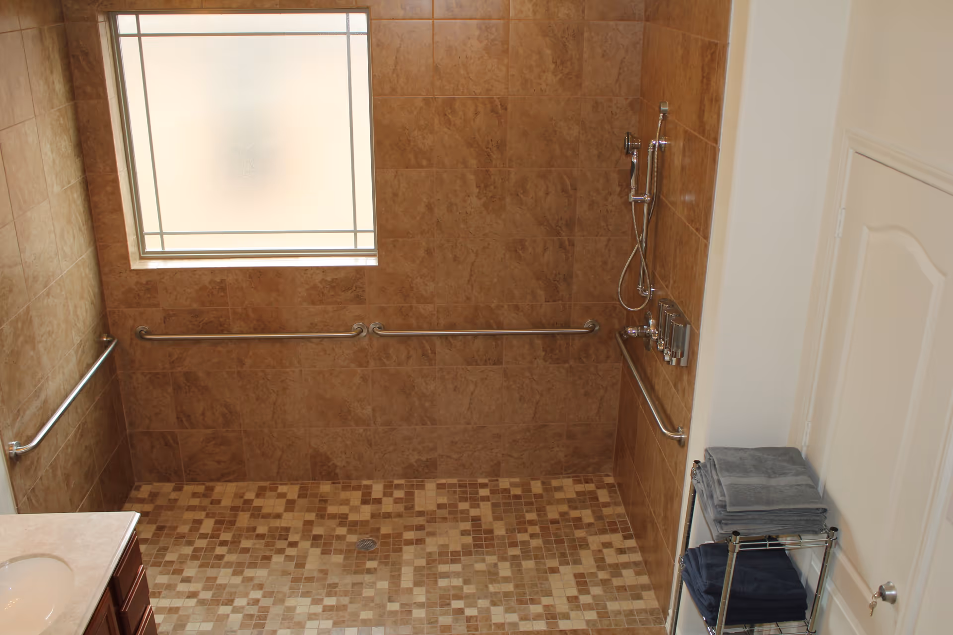 A spacious walk-in shower with beige and brown tiled walls and floor, equipped with multiple grab bars and a handheld showerhead. There is a frosted window providing natural light, a white door to the right, and a metal rack holding folded towels.