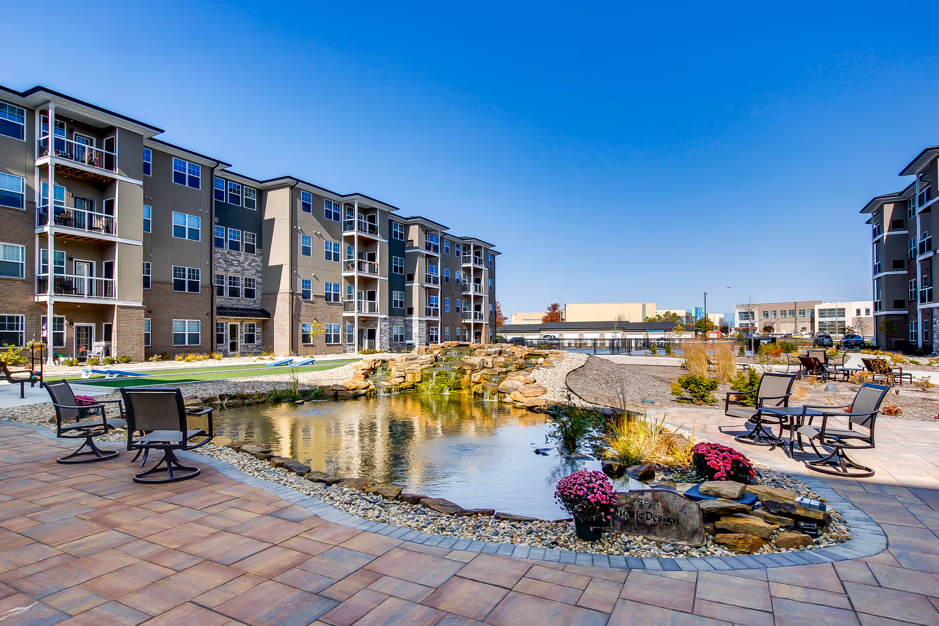 Outdoor courtyard of a modern apartment complex with a pond, patio seating, and multi-story buildings under a clear blue sky.