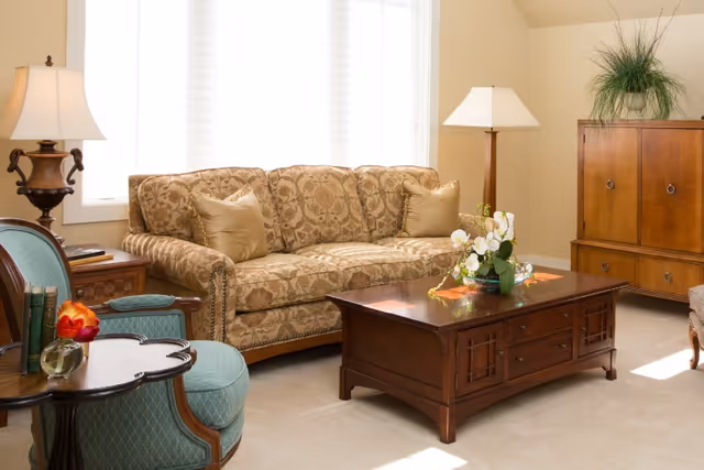 A well-lit living room with a patterned sofa, wooden coffee table, armchairs, lamps, and a wooden cabinet.