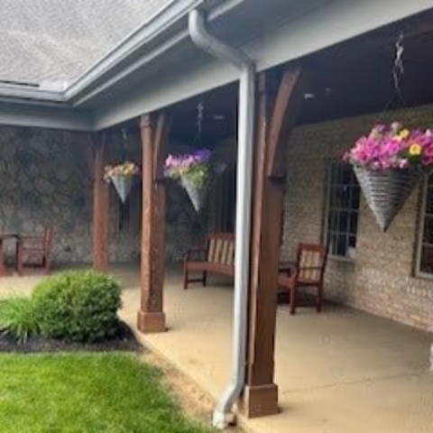 Covered outdoor patio area with wooden posts and hanging flower baskets. There are benches along the brick wall and a small green bush near the edge of the patio. The roof has a gutter and downspout visible.