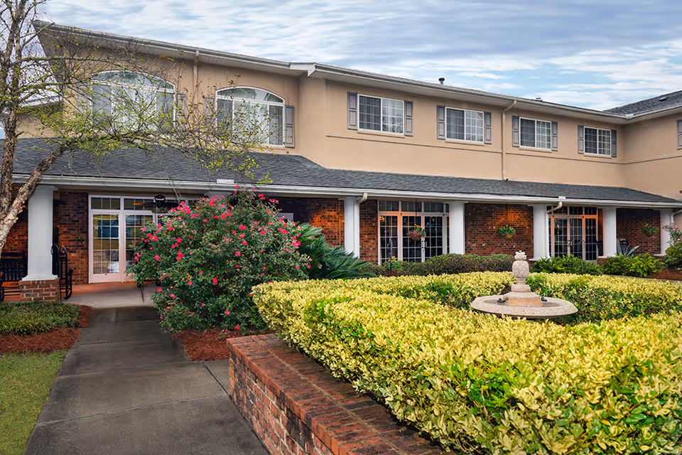Front entrance of a two-story senior living facility with a landscaped courtyard, hedges and a small fountain.