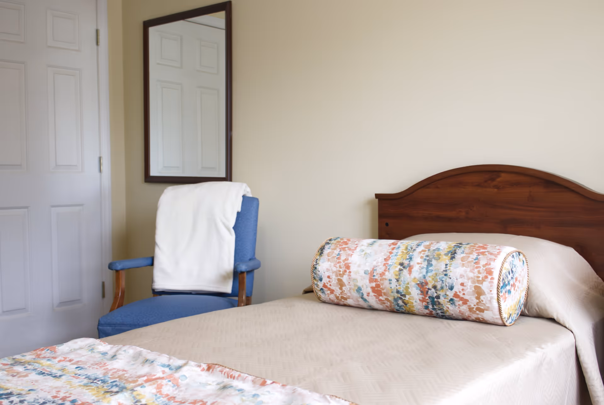 A neatly made bed with a wooden headboard and a colorful cylindrical pillow on top. Next to the bed is a blue upholstered chair with wooden armrests, draped with a white blanket. A rectangular mirror hangs on the wall above the chair, reflecting a closed white door.