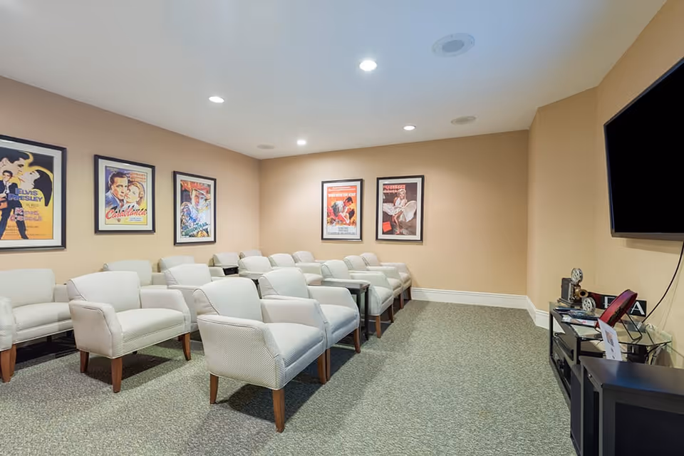 A small theater room with two rows of beige upholstered armchairs facing a large flat-screen TV mounted on the wall. The walls are beige and decorated with framed classic movie posters including Elvis Presley and Casablanca. The room has recessed ceiling lights and a carpeted floor.