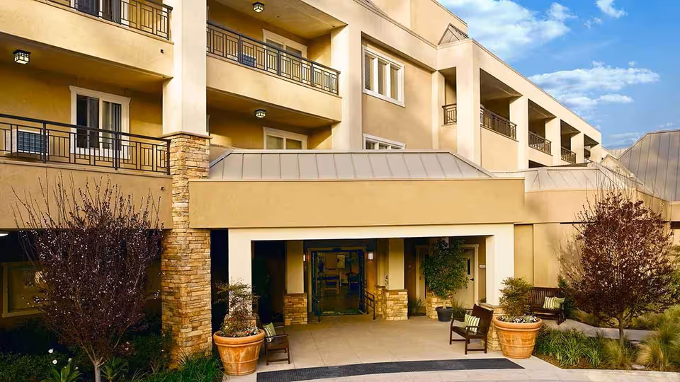 Exterior front entrance of a multi-story senior living building with balconies, a covered entry, potted plants and benches.