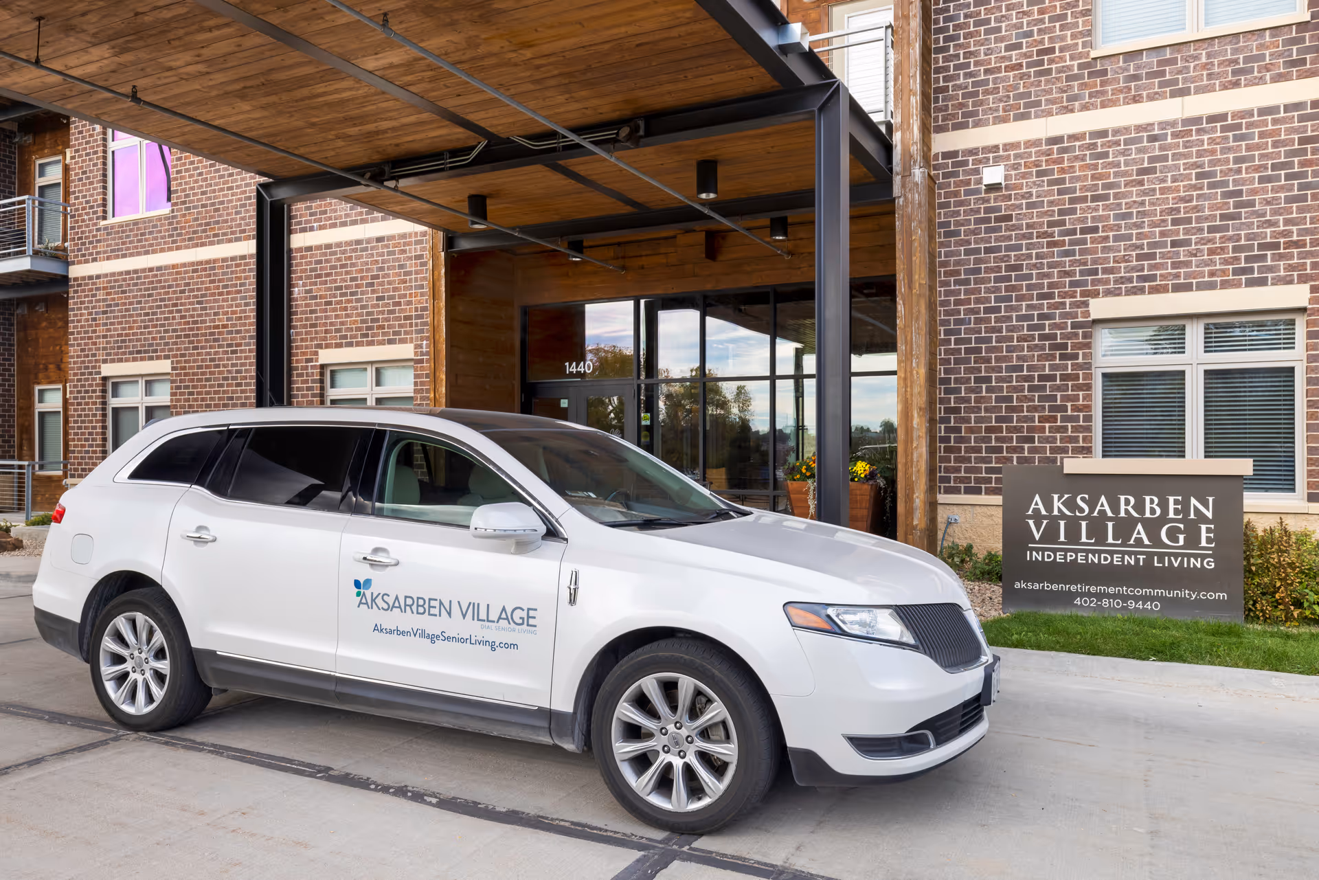 A white SUV with Aksarben Village branding parked in front of the entrance to a brick building with a covered driveway. A sign next to the entrance reads 'Aksarben Village Independent Living' along with the website and phone number.