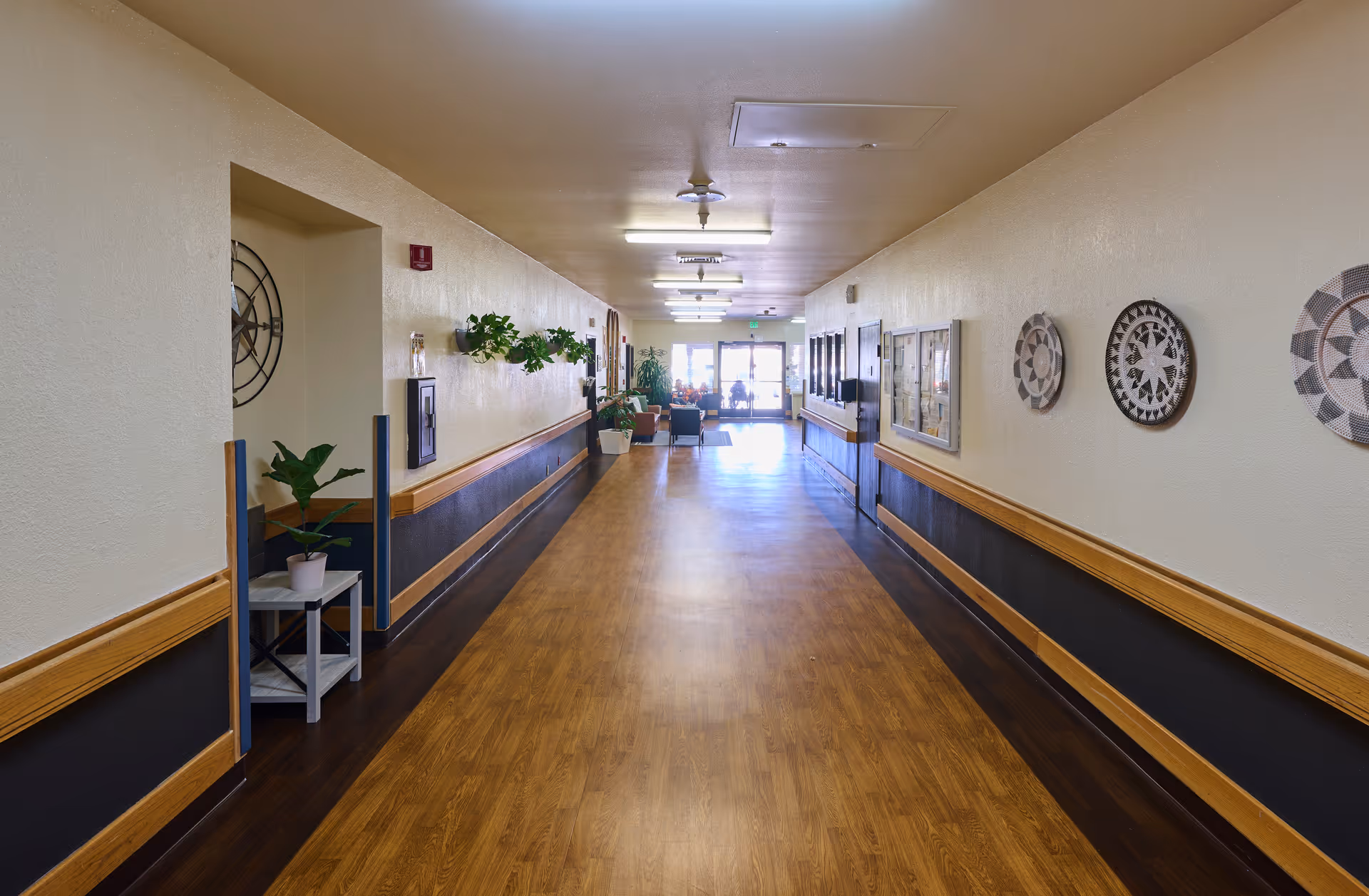 Long, well-lit interior hallway of a nursing facility with wood flooring, handrails, wall decor, potted plants, and seating visible at the far end.