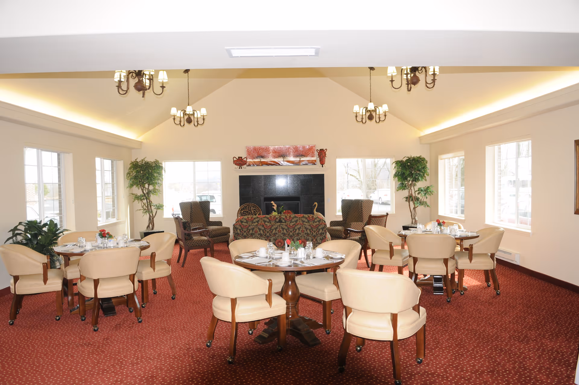 A bright and spacious dining room with multiple round tables set with white dishes, glasses, and silverware. Beige upholstered chairs surround the tables. The room has large windows letting in natural light, red patterned carpet, and cream-colored walls. There are two tall green plants near the windows and a fireplace with a decorative painting above it. Several chandeliers hang from the vaulted ceiling.