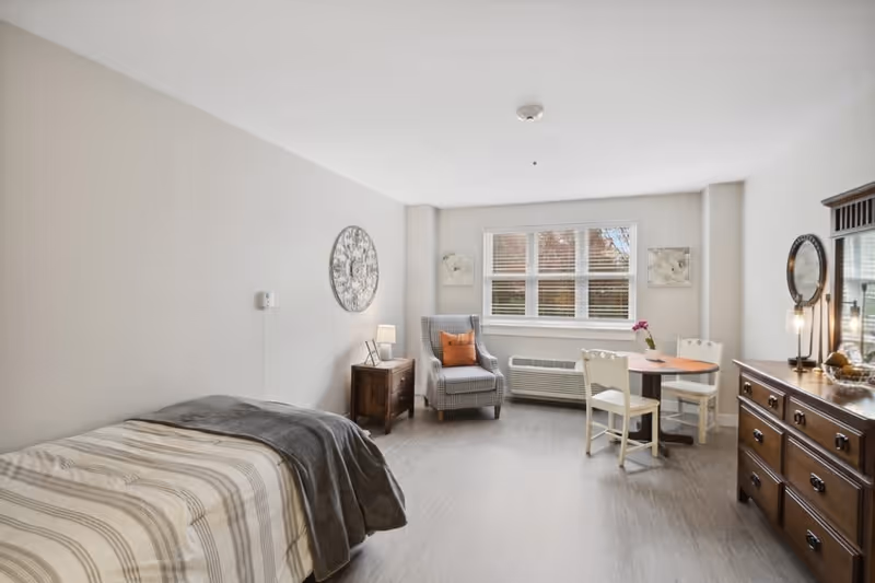 A bright and neatly arranged bedroom with a single bed covered in striped bedding and a gray blanket. Next to the bed is a wooden nightstand with a lamp and a decorative clock on the wall above. Across from the bed is a wooden dresser with a mirror and two lamps. A small round table with two chairs and a flower vase is positioned near a window with blinds. A comfortable armchair with an orange cushion is placed near the window, and the room has light-colored walls and flooring.