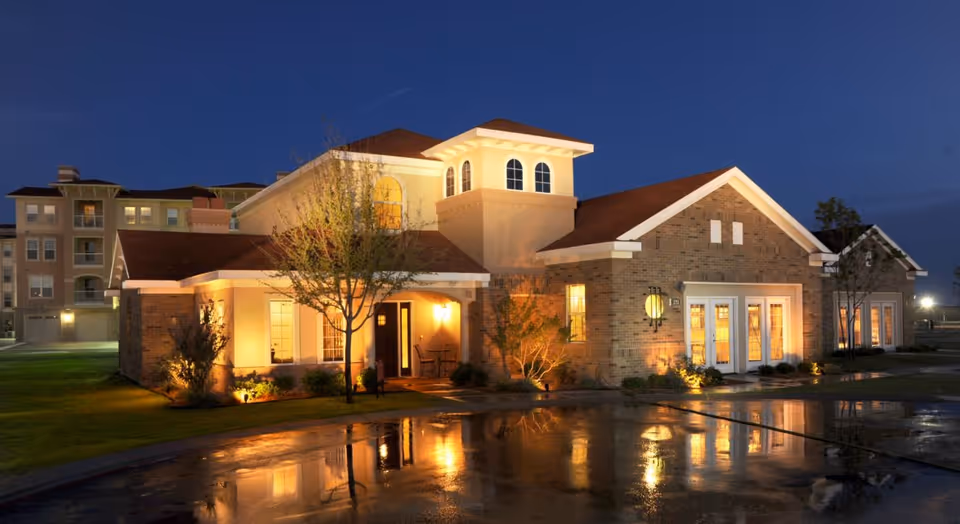 Exterior view of a well-lit brick building at dusk with reflections on a wet driveway, surrounded by grass and trees, with a multi-story building visible in the background.