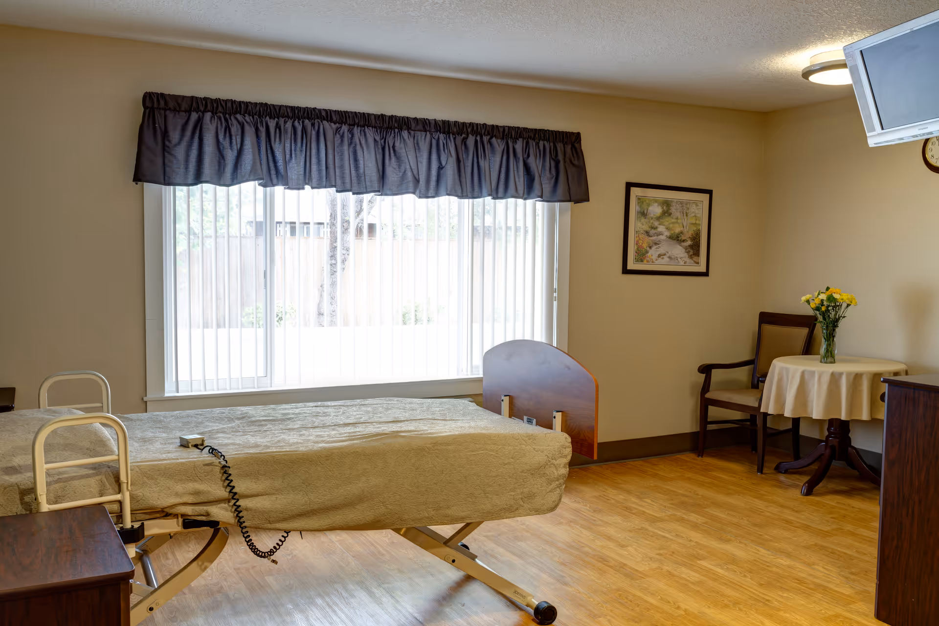 A senior living facility bedroom with a single adjustable bed covered in a beige blanket, a wooden nightstand, a window with vertical blinds and a dark valance, a small round table with a vase of yellow flowers, a wooden chair, a wall-mounted TV, and a framed landscape painting on the wall.