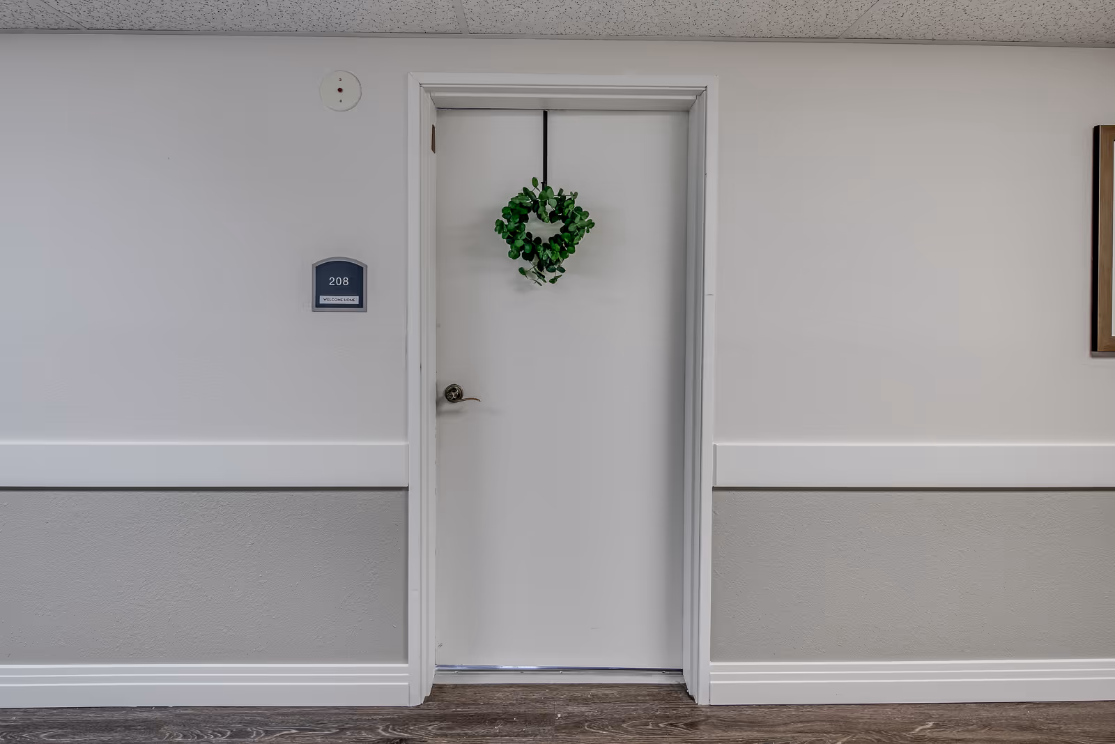 A white door in a hallway with a green wreath hanging on it. The door has a silver handle and a room number plaque to the left that reads '208'. The walls are painted white and gray with a chair rail, and the floor is dark wood. Part of a framed picture is visible on the right side of the image.