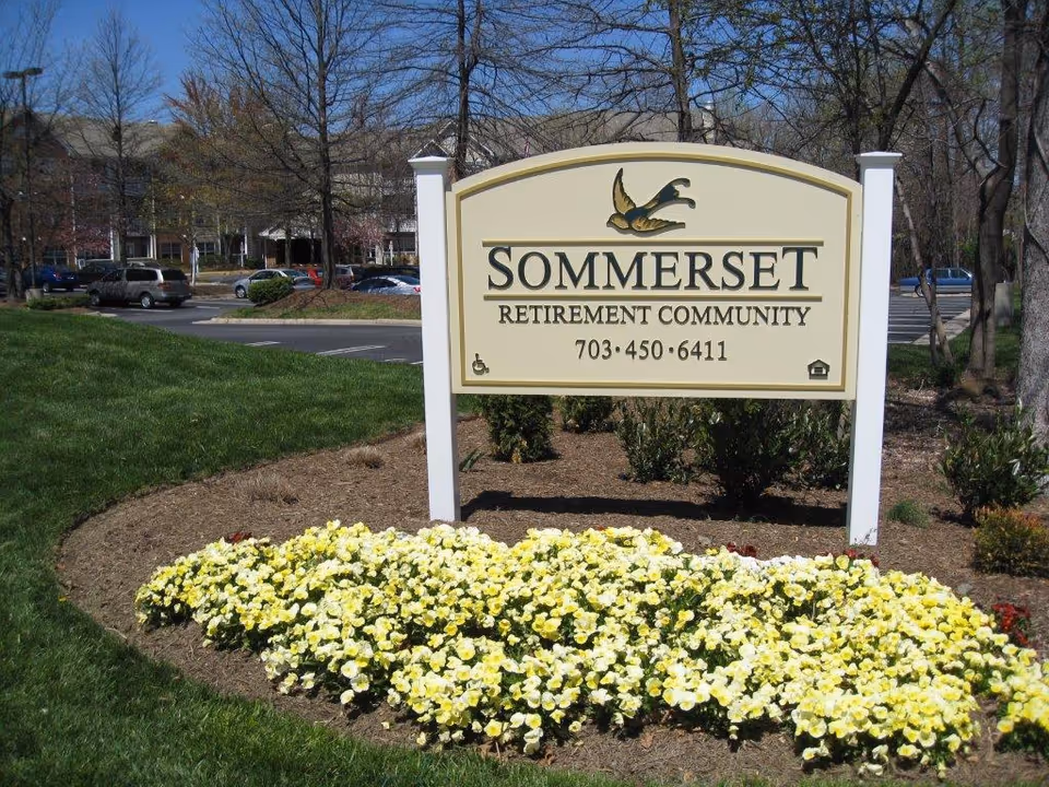 Outdoor view of a sign for Sommerset Retirement Community with a phone number 703-450-6411, surrounded by a flower bed with yellow and white flowers, grass, trees, and parked cars in the background.