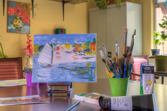 A painting of a sailboat on water with colorful trees in the background displayed on a small easel on a table. The table also holds a green container filled with paintbrushes, a tube of acrylic paint, and other art supplies. The room has yellow walls, a cabinet, and some plants near a window letting in natural light.
