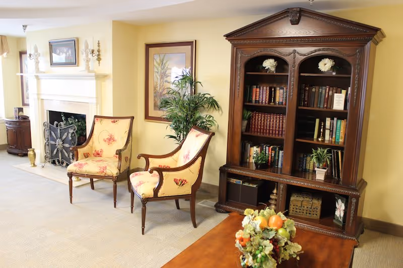 A cozy living room area with two floral upholstered armchairs, a wooden coffee table with a floral centerpiece, a large wooden bookshelf filled with books and decorative plants, a fireplace with a decorative screen, and framed artwork on the walls.