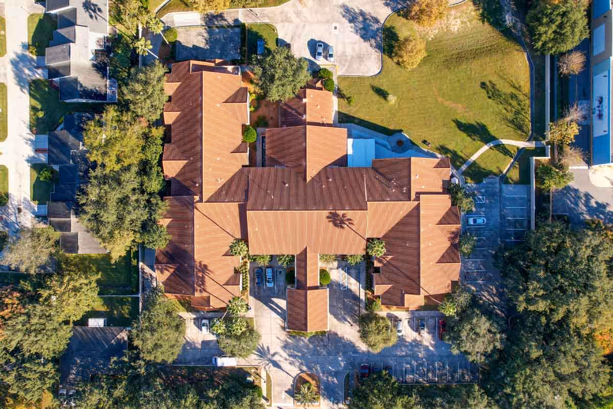 Aerial view of a large building with a red-tiled roof surrounded by trees, parking lots, and green grassy areas, showing the layout of the facility and its surroundings.
