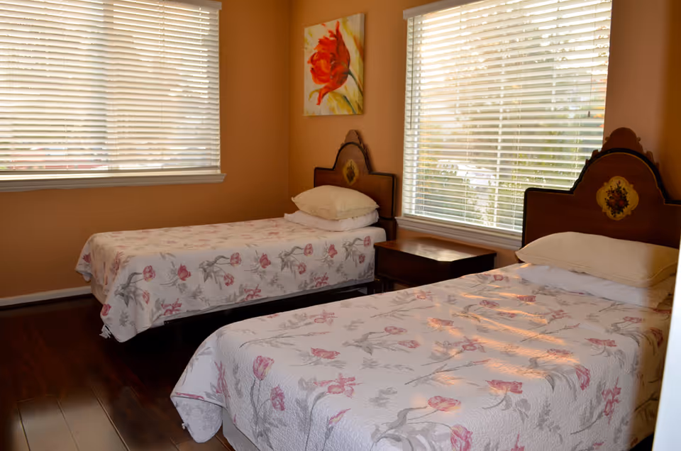 Sunlit bedroom with two single beds in floral quilts, wooden headboards, a nightstand between them, and large windows with blinds.