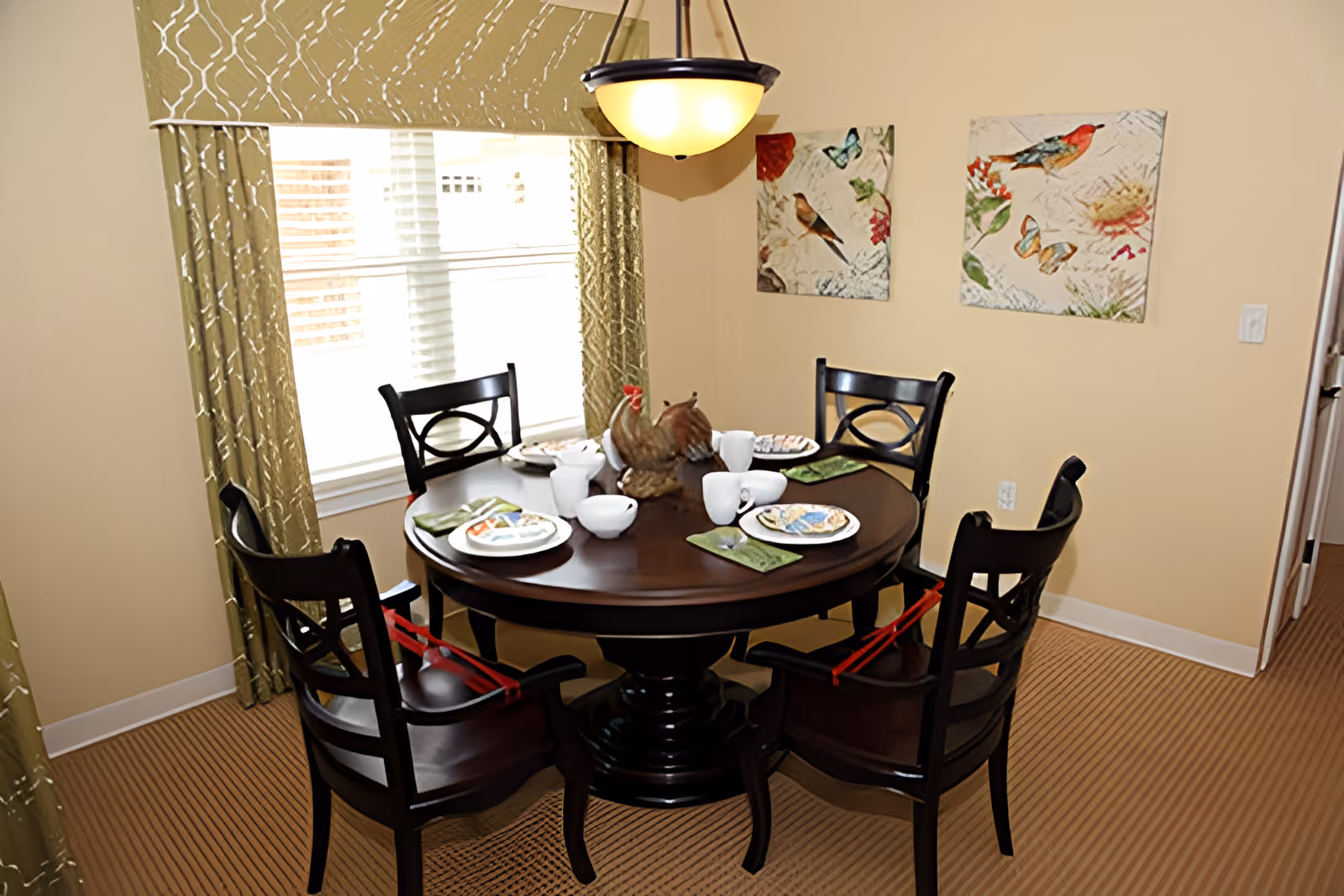 A dining room with a round dark wooden table set for four people. The table has plates, bowls, cups, and green napkins, with a decorative rooster centerpiece. Four dark wooden chairs with armrests surround the table, each with a red strap attached. The room has beige walls, a window with green patterned curtains, and two bird-themed paintings on the wall. A hanging light fixture is above the table.