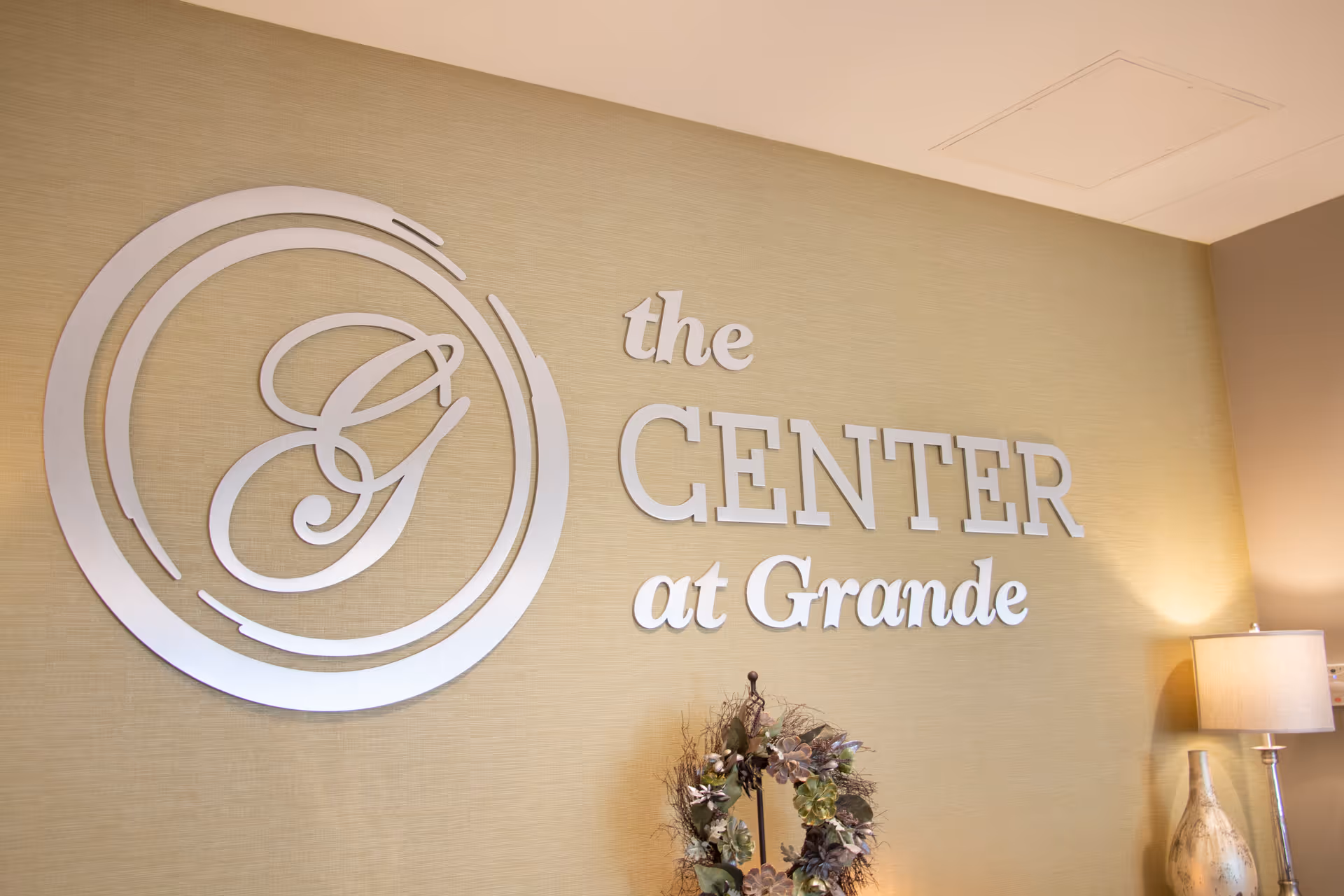 Wall with a decorative sign that reads 'the CENTER at Grande' in silver letters, accompanied by a circular logo with the letter G. Below the sign is a floral wreath, and to the right is a table lamp with a beige shade.