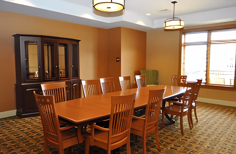 A dining room with a long wooden table surrounded by wooden chairs. There is a dark wooden cabinet against the wall and two ceiling lights hanging above the table. The room has beige walls, a patterned carpet, and a large window letting in natural light.