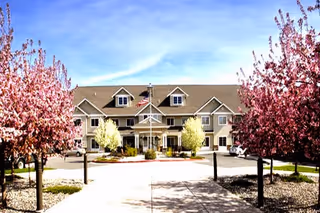 Front exterior view of a senior living facility building with a peaked roof, multiple windows, and an American flag on a flagpole in front. The entrance is framed by trees with pink blossoms on either side of the walkway leading to the building.