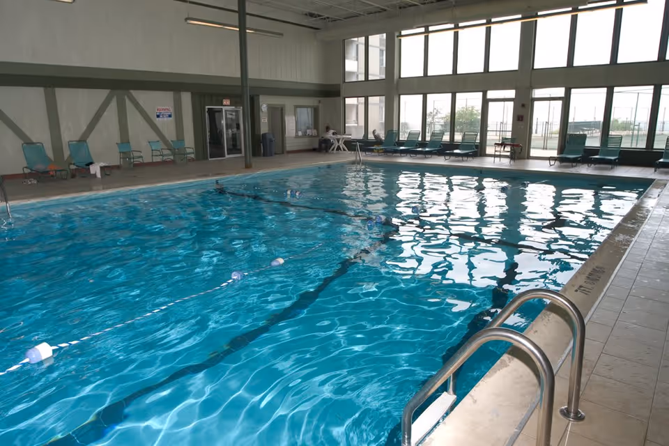 Indoor swimming pool with clear blue water, lane dividers, and metal handrails. Several green lounge chairs are arranged along the poolside, with large windows letting in natural light. A person is seated at a table near the back wall, and there are doors and signs visible on the far wall.
