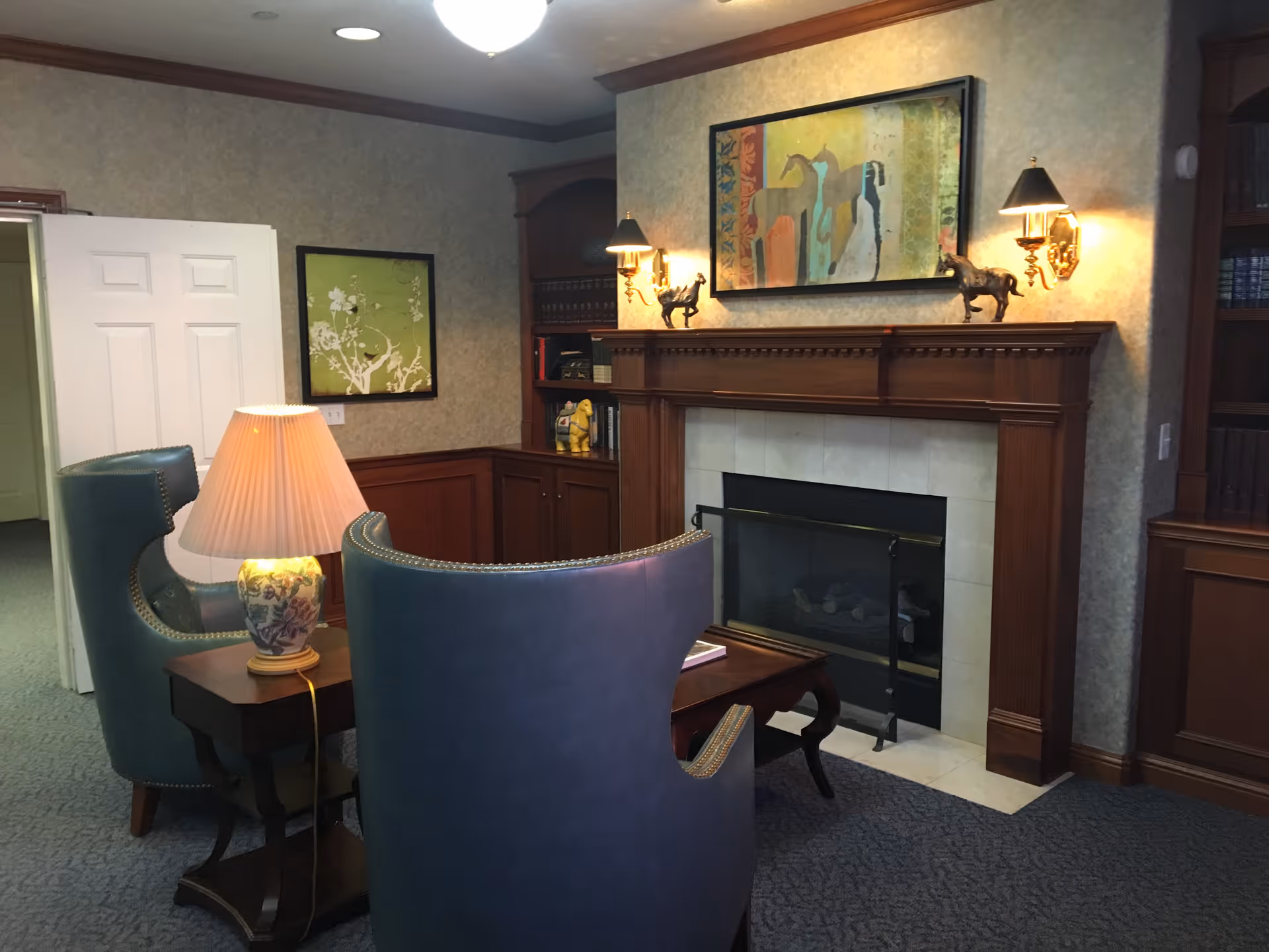 A cozy interior room with two blue upholstered armchairs facing a wooden fireplace mantel. The mantel is decorated with two horse figurines and a colorful painting of horses above it. There are wall-mounted lamps on either side of the painting, a table lamp on a small wooden side table between the chairs, and built-in wooden bookshelves with books and decorative items. The room has carpeted flooring and a partially open white door leading to another area.