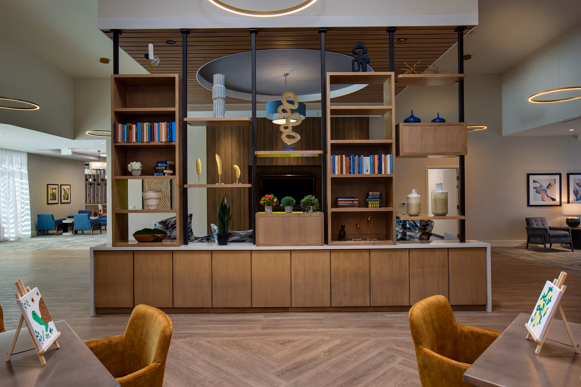 Open communal lounge with a central wooden shelving divider, seating, decorative objects, and small art easels on tables.