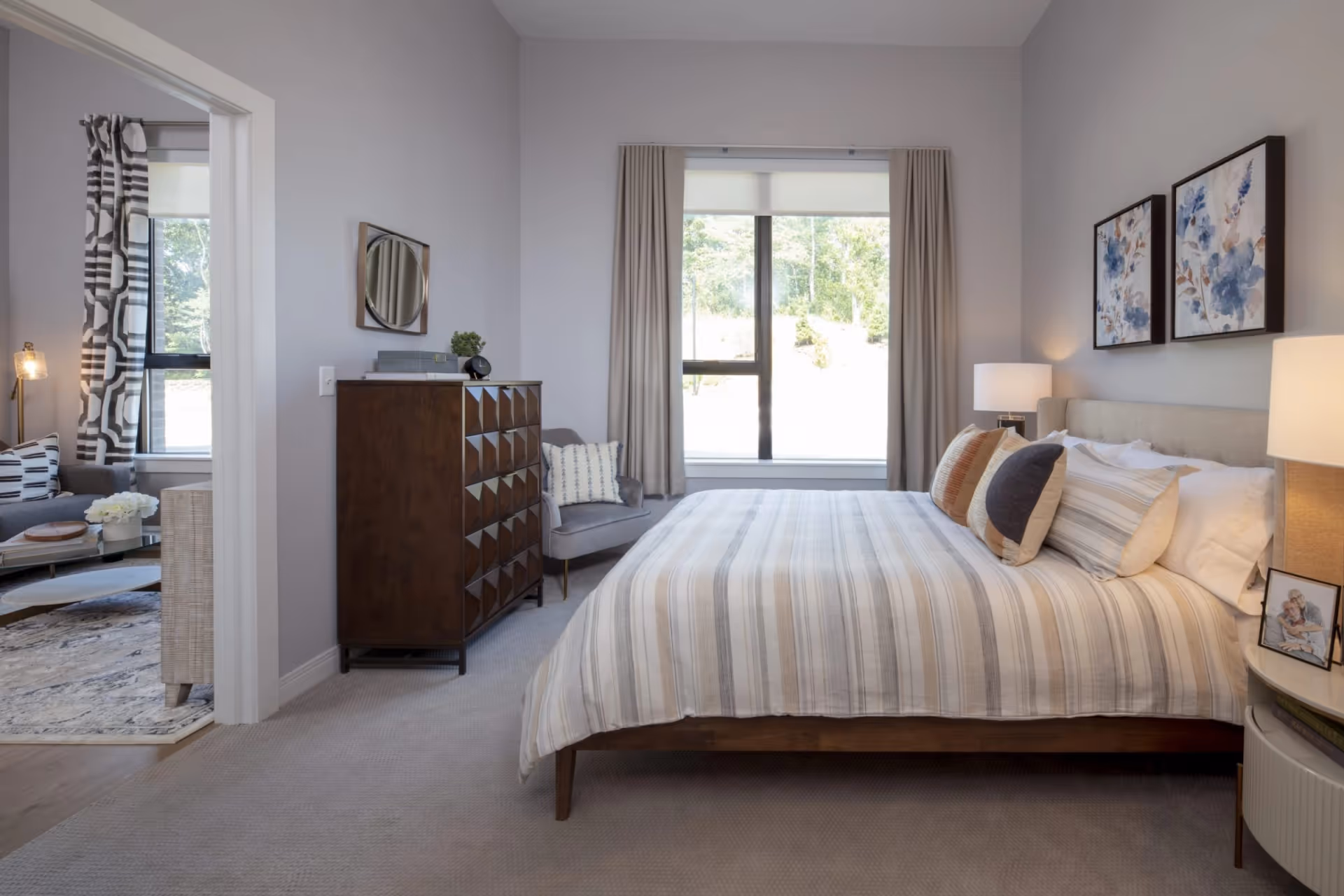 Well-lit bedroom featuring a striped bed with accent pillows, bedside lamps, a wooden dresser, an armchair, and a large window overlooking greenery.