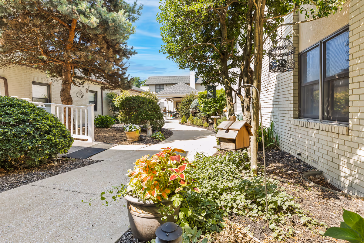 A sunny outdoor walkway at a senior living facility with well-maintained landscaping including bushes, trees, and potted plants. There is a birdhouse hanging from a tree on the right side near a beige brick building with windows. The path curves gently and leads to more buildings in the background under a clear blue sky.