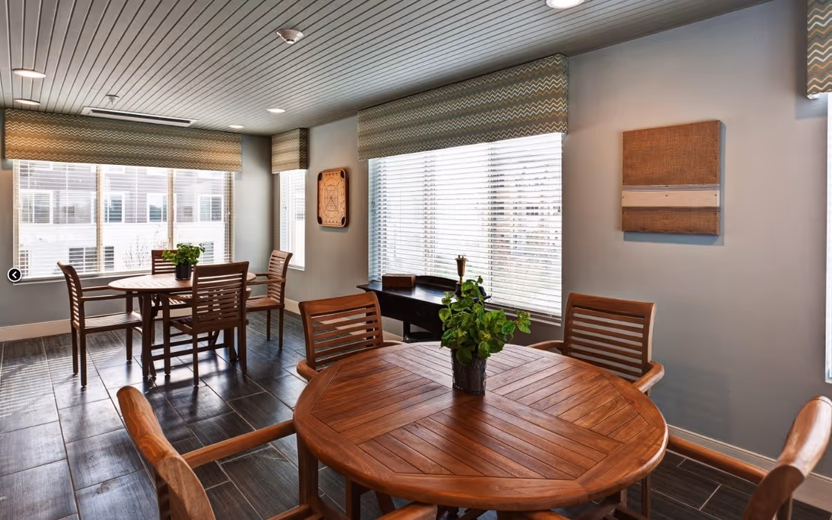 A bright and clean interior room with two wooden round tables, each surrounded by wooden chairs. The room has large windows with blinds and patterned valances, letting in natural light. There are two pieces of wall art and a small desk with decorative items near the windows.