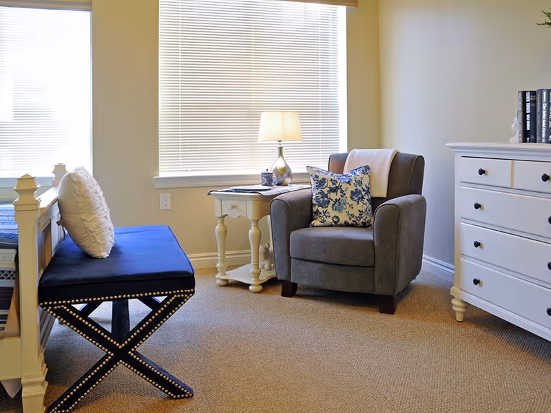 A cozy sitting area in a senior living facility room featuring a gray armchair with a floral cushion and a light blanket draped over the back, a small white side table with a lamp and decorative items, a blue cushioned bench with a white pillow, and a white dresser with black knobs. The room has beige carpet and large windows with blinds letting in natural light.