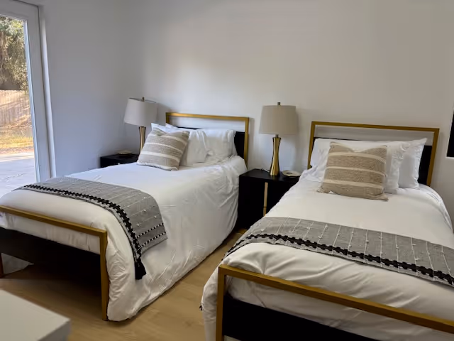 Two neatly made twin beds with white linens, decorative pillows and throw blankets in a bright bedroom with bedside tables and lamps.