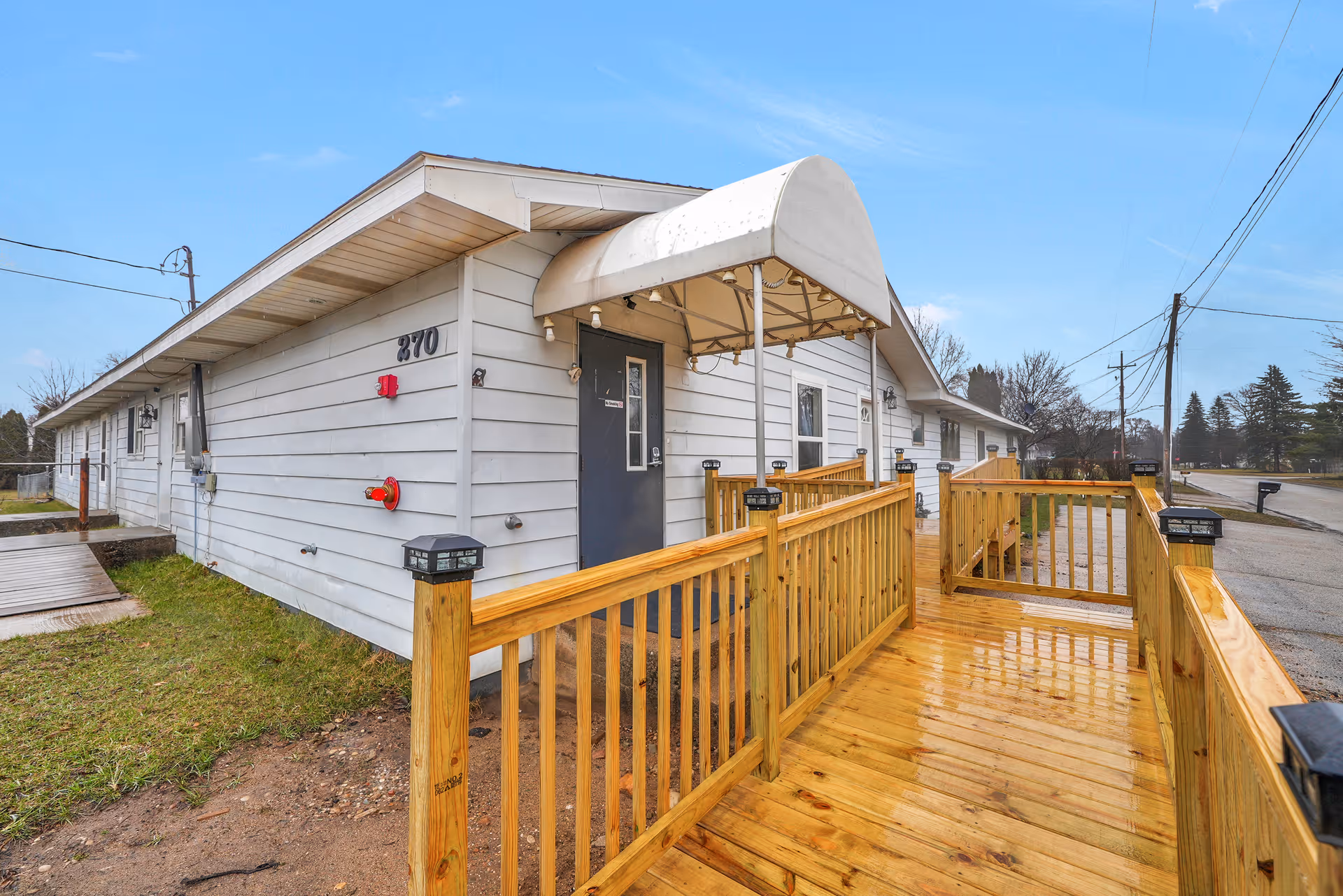Exterior view of a single-story white building with a wooden ramp leading to a door under a white awning. The building has the number 270 on the side and is situated next to a street with utility poles and trees in the background.