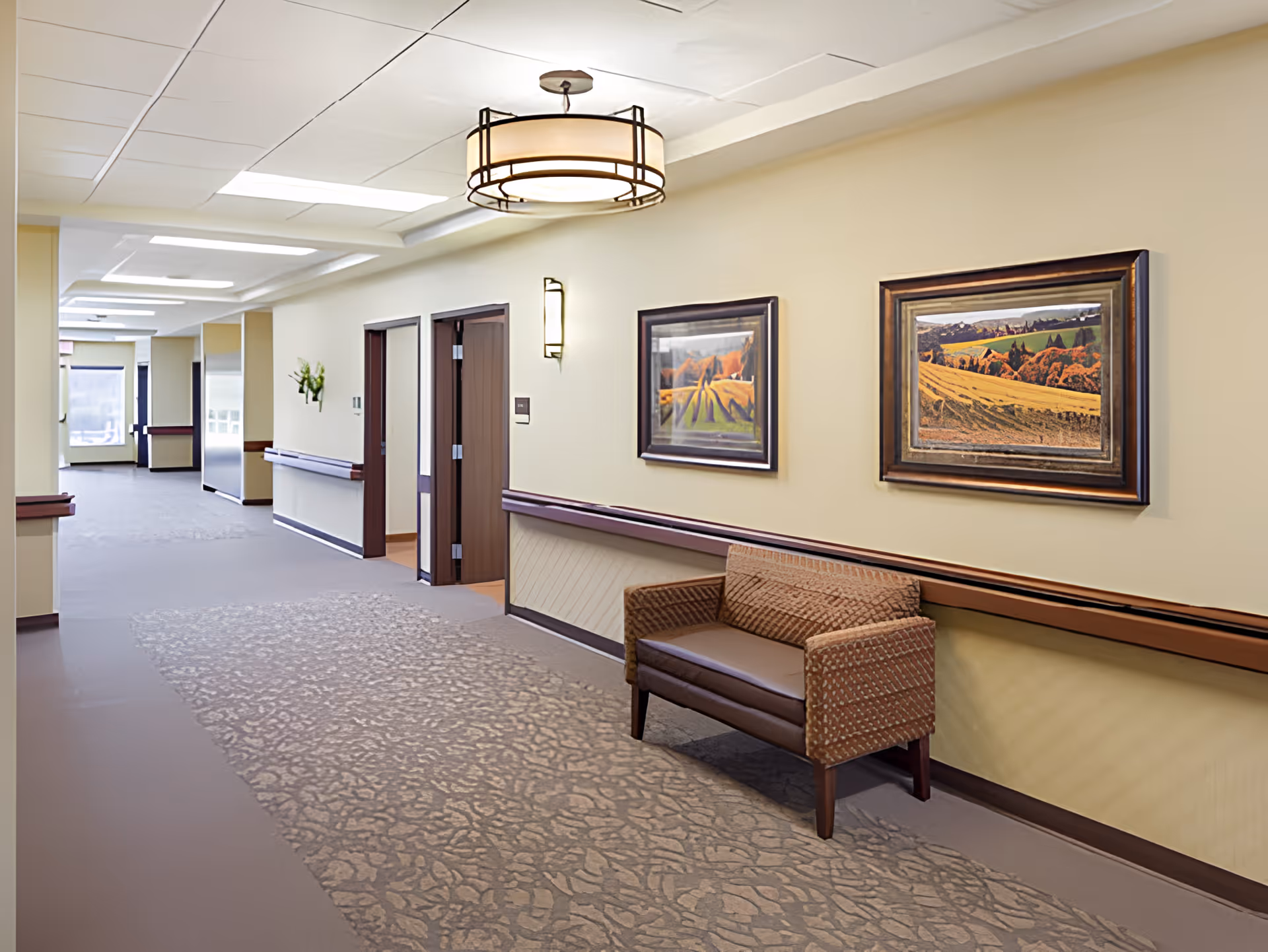 A well-lit hallway in a senior living facility with beige walls and carpeted floor. The hallway features a brown cushioned bench, two framed landscape paintings on the wall, wooden handrails, and ceiling lights. Several doorways and a window are visible further down the corridor.