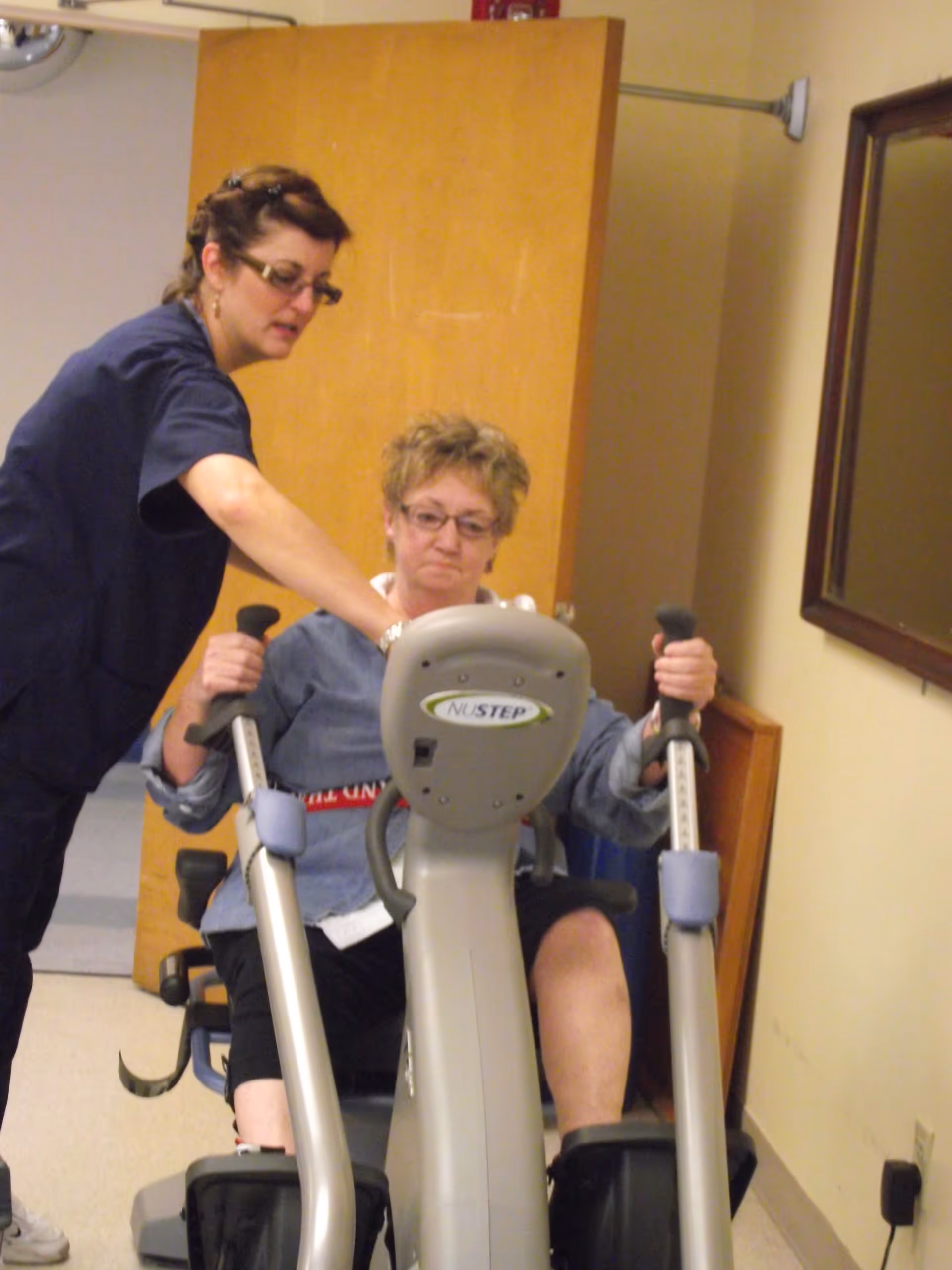 A woman seated on a NuStep exercise machine is being assisted by a healthcare worker in a room with beige walls and a wooden door. The healthcare worker is pointing at the machine's controls while the seated woman holds the handles.