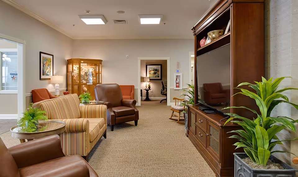 A cozy living room area in a senior living facility featuring a large wooden entertainment center with a flat-screen TV, several comfortable chairs and a striped sofa, side tables with green plants, a wooden rocking chair, and a glass display cabinet against the far wall. The room is well-lit with ceiling lights and decorated with framed artwork and neutral-colored walls and carpet.