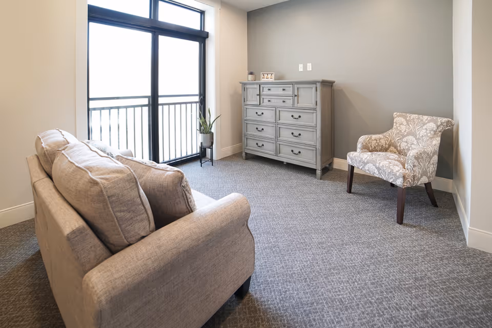 A cozy living room area with a beige sofa, a patterned armchair, a gray dresser, and a large window with a black frame letting in natural light. There is a small potted plant on a stand near the window.