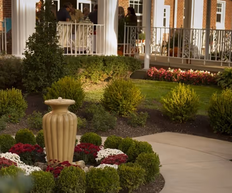 Landscaped courtyard with a small stone fountain, trimmed shrubs and flowerbeds in front of a porch where several people are standing.