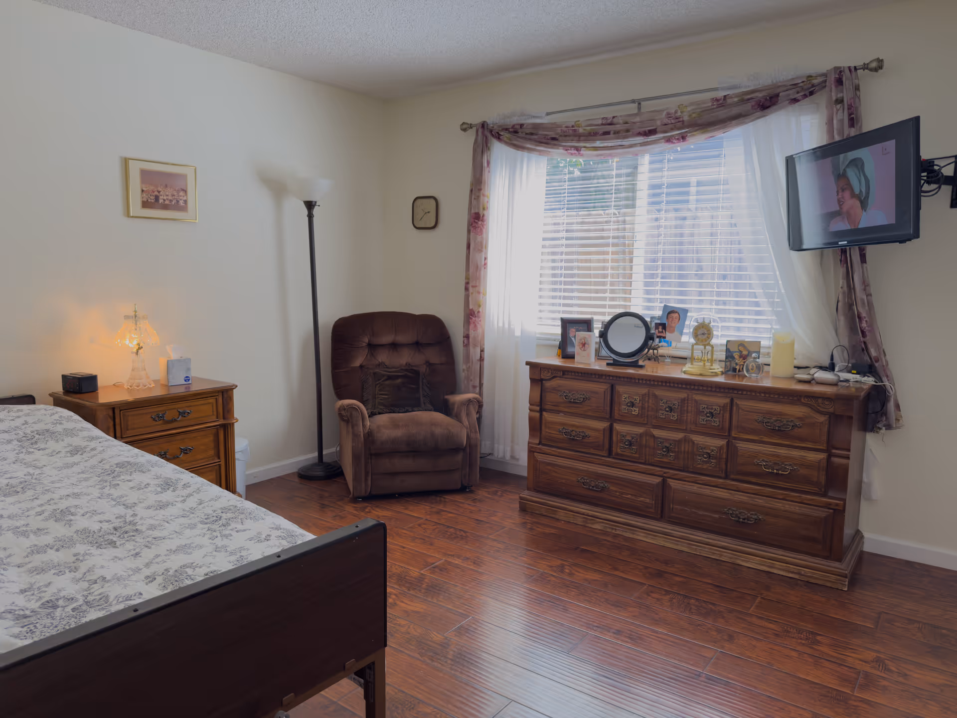 A cozy bedroom in Citrus Garden Care Home featuring a bed with patterned bedding, a wooden nightstand with a lamp and tissue box, a brown recliner chair, a wooden dresser with framed photos and decorative items, a wall-mounted TV, and a window with floral curtains letting in natural light.