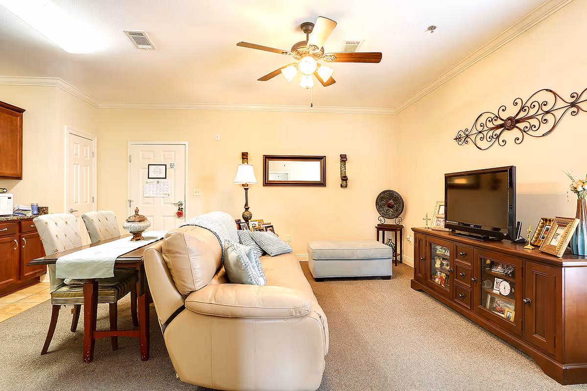 A cozy senior living room featuring a beige leather sofa with cushions, a wooden TV stand with a flat-screen television, decorative wall art, a ceiling fan with lights, a small ottoman, and a dining table with upholstered chairs adjacent to a kitchen area.