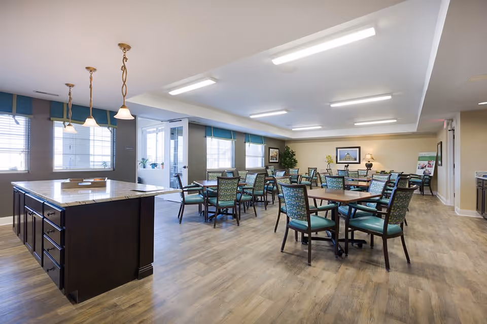 A spacious dining room with multiple tables and chairs arranged neatly. The room features large windows with blue valances, a central island with a marble countertop, pendant lights hanging above it, and framed artwork on the walls. The flooring is wood-style, and the room is well-lit with ceiling lights.