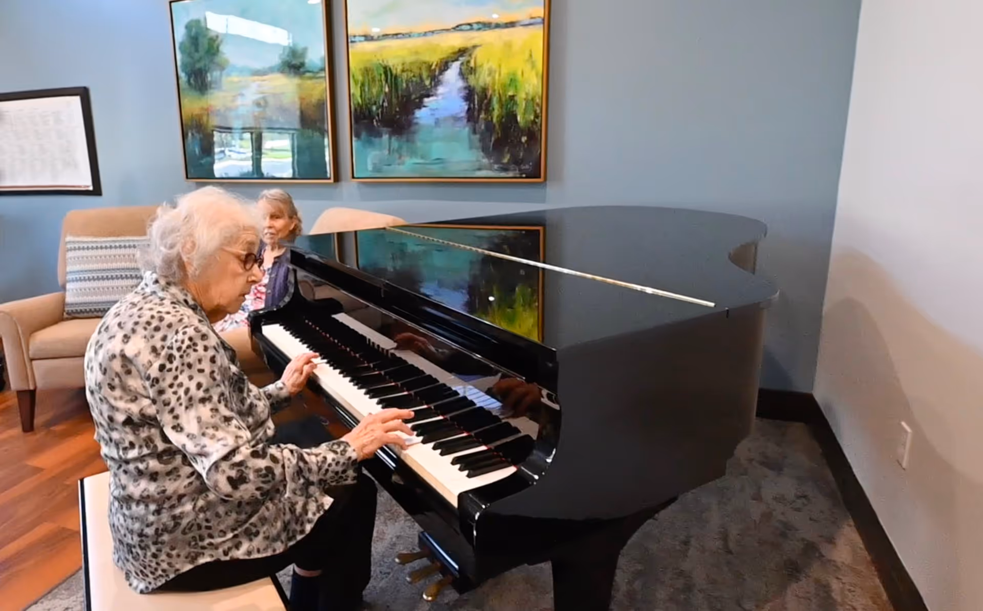 An elderly woman with white hair and glasses is playing a black grand piano in a room with blue walls. Another elderly woman is seated on a beige armchair in the background. Two framed landscape paintings hang on the wall behind them.
