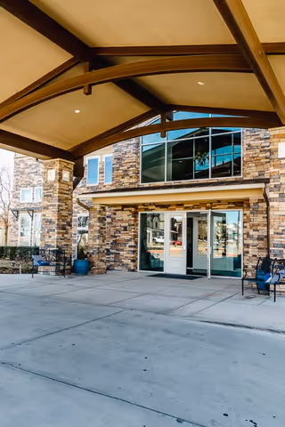 Entrance of a senior living facility with a covered drop-off area, stone facade, large glass doors, and benches on either side.
