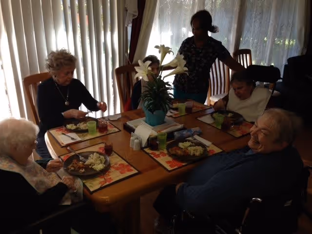 A group of elderly people sitting around a dining table having a meal together in a room with vertical blinds and a vase with white lilies in the center of the table. A caregiver is standing behind them assisting.
