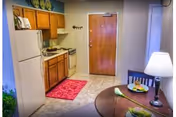 Interior of a small studio showing a kitchenette with wood cabinets and white appliances, an entry door, and a round dining table with a lamp.