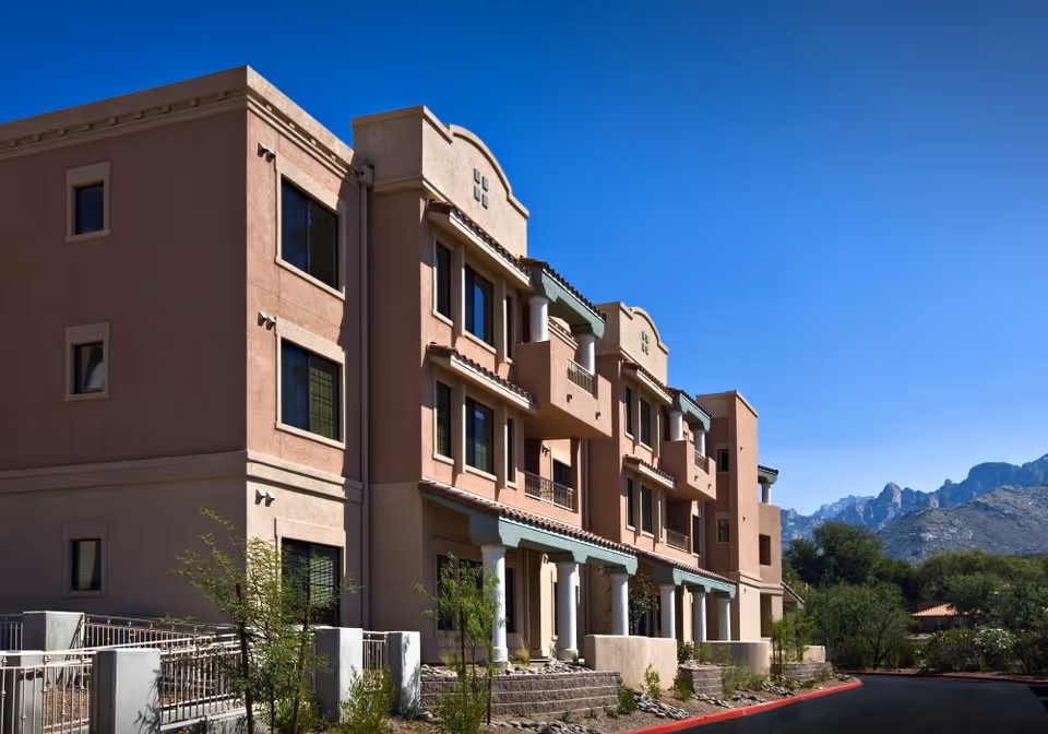Exterior view of a multi-story senior living facility building with balconies and columns, set against a clear blue sky and mountainous background.