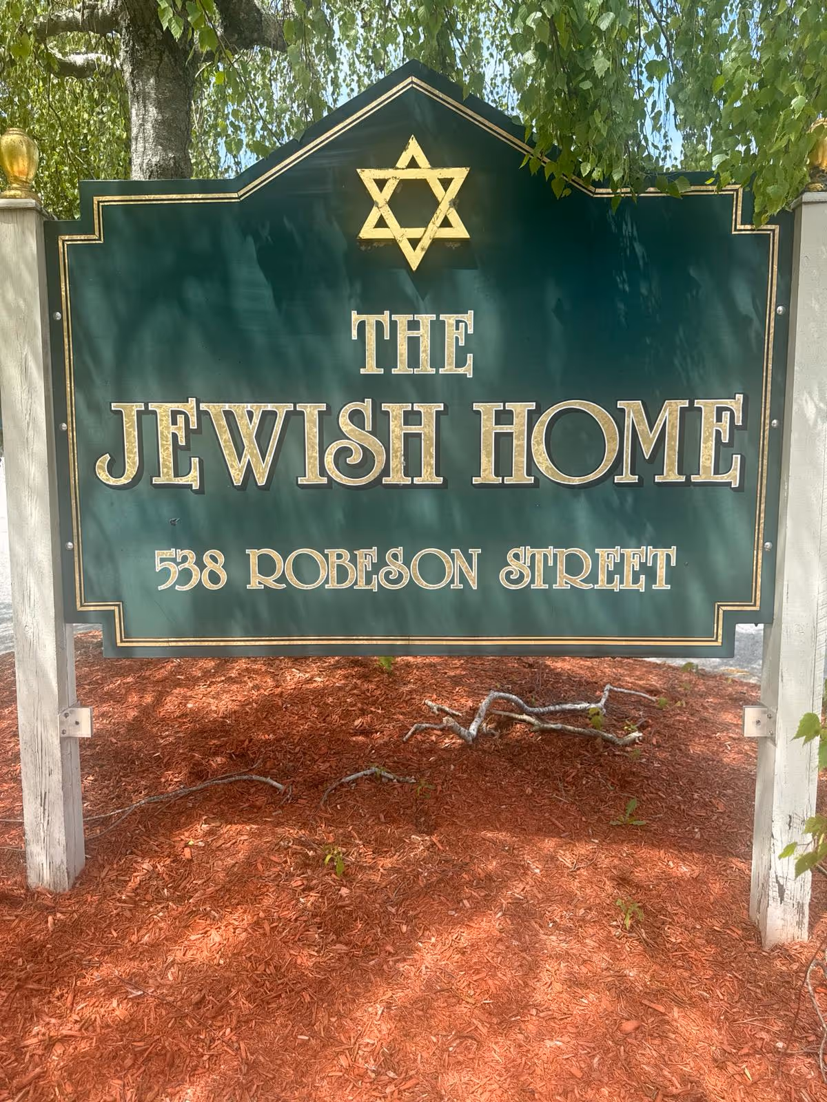 Green outdoor sign reading "THE JEWISH HOME 538 ROBESON STREET" with a Star of David mounted above red mulch under a tree.