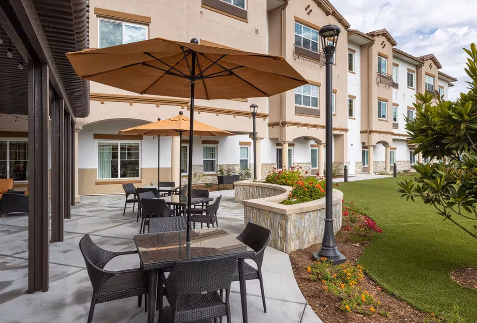 Outdoor courtyard patio with tables, umbrellas and chairs beside a landscaped lawn in front of a three-story senior living building.