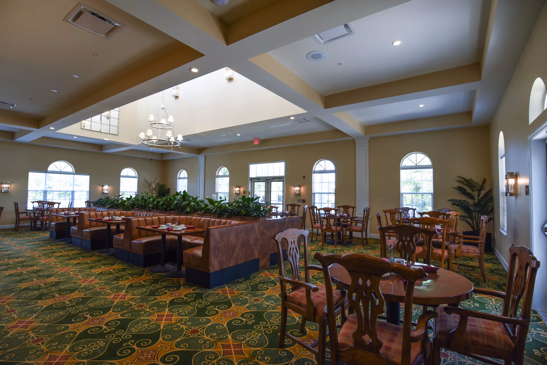 Spacious dining room with multiple wooden tables and chairs, booth seating with brown leather upholstery, green patterned carpet, large windows with arched tops, and a chandelier hanging from a high ceiling with recessed lighting.